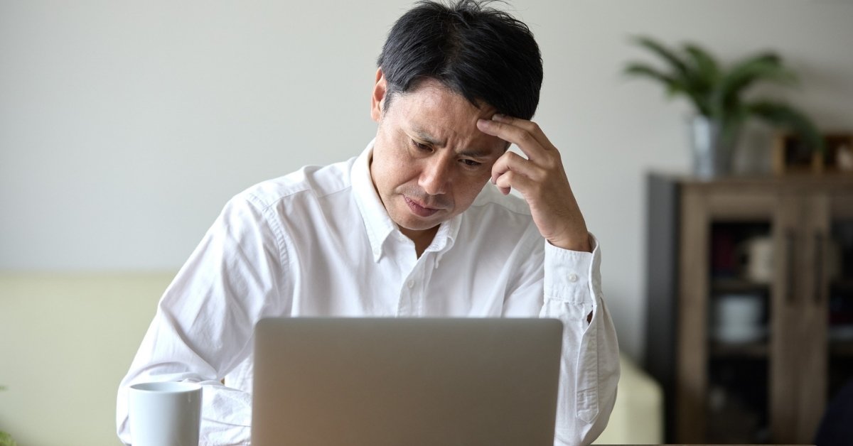 A man in a white shirt sitting at a laptop with a concerned expression, resting his head on his hand.
